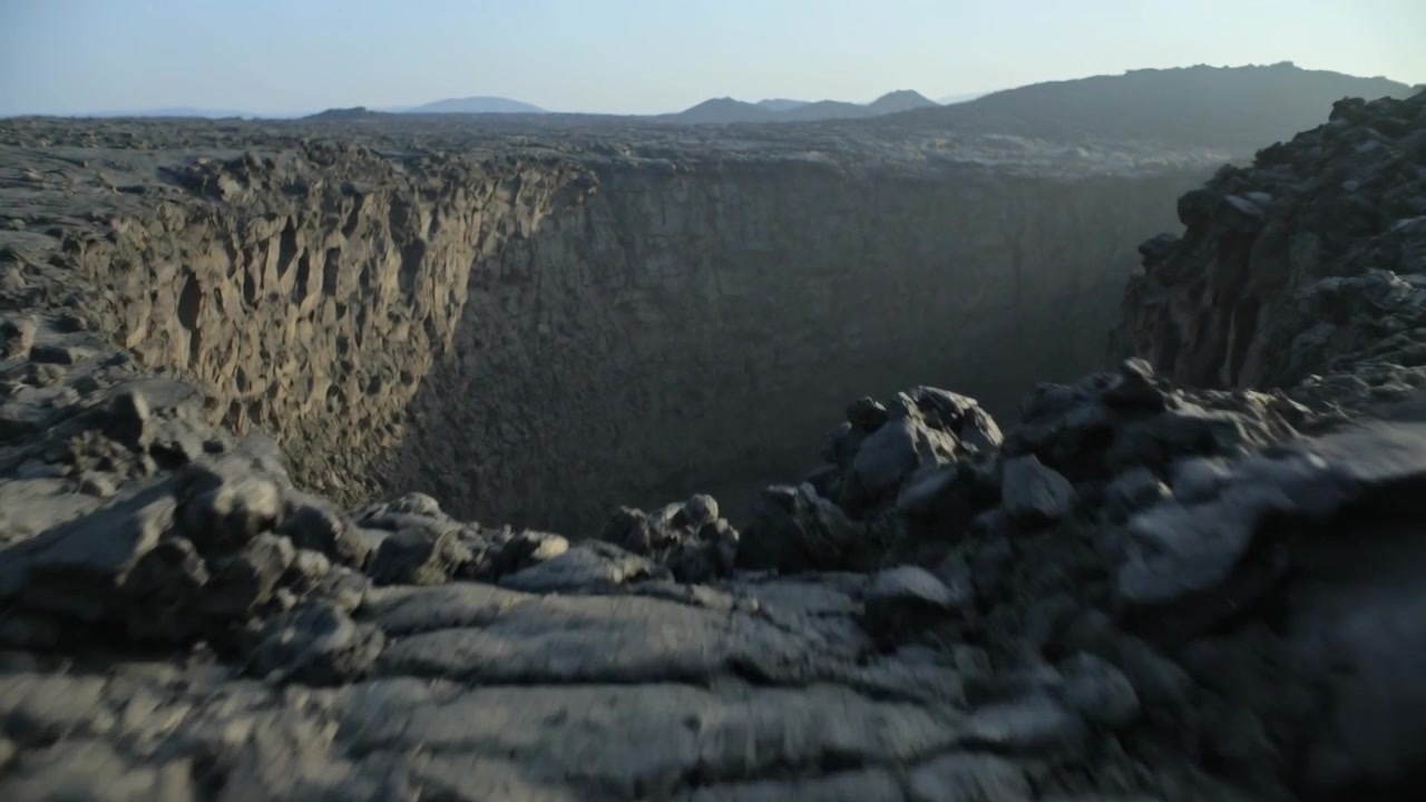 Kameraet beveger seg fremover og avslører et kollapskrater mens det beveger seg over et lavafelt, for å avsløre klippevegger