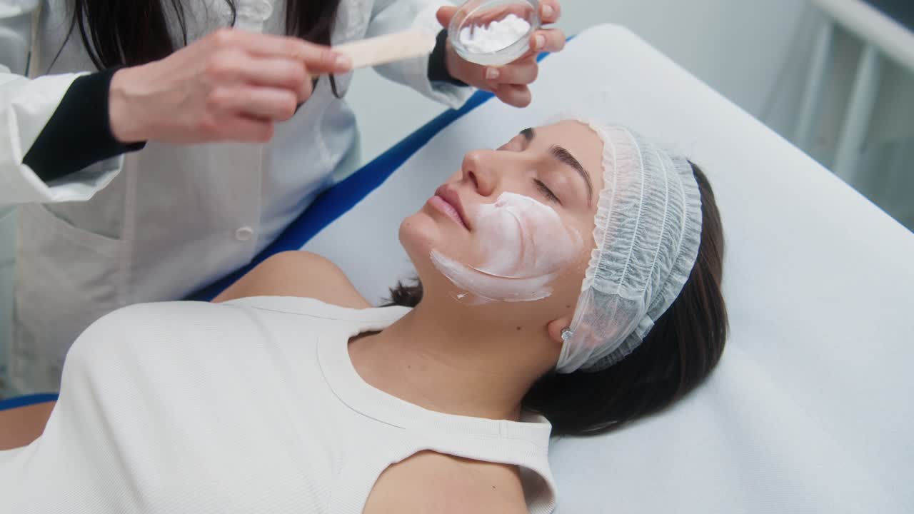 Estetista usa una spatola per stendere una maschera in crema bianca sul viso del cliente durante una sessione di trattamento skincare in spa