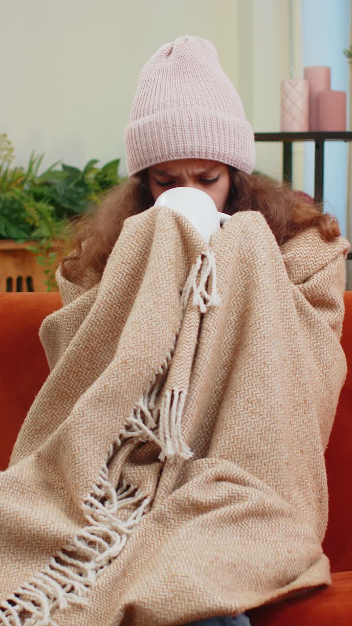 Sick teen girl kid wear hat wrapped in plaid sit alone shivering from cold on sofa drinking hot tea