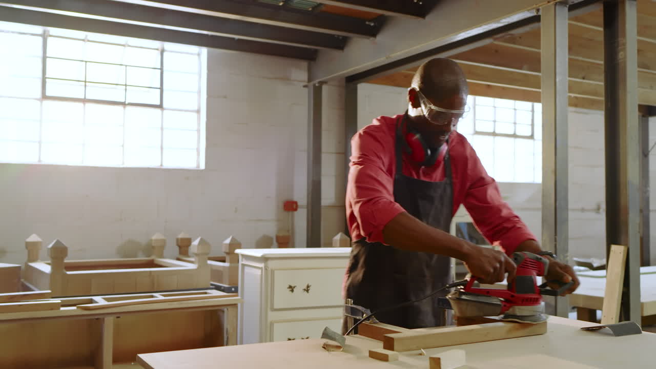 African American male carpenter picking up sander, sanding and finishing wood board at workbench