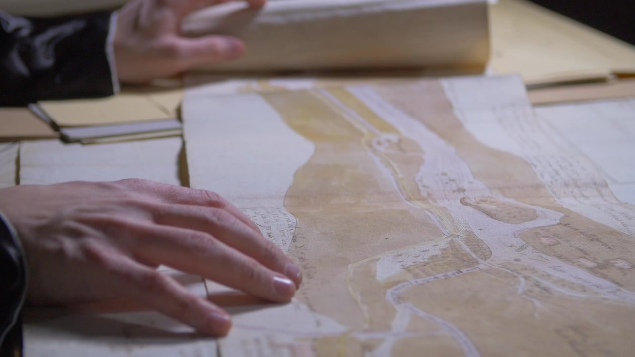 Nærbillede af hænder, der forsigtigt åbner et antikt kort på et træbord. Koncept for historisk kartografi, udforskning, geografi, kulturarv og arkivforskning