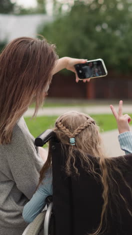 mother makes fun selfies with little daughter in wheelchair sitting in summer park. girl shows finger gestures in camera of smartphone backside view