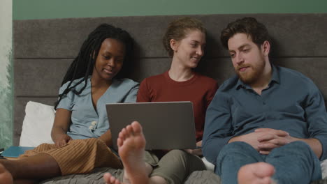 three roommates are watching videos on laptop sitting on the bed in bedroom