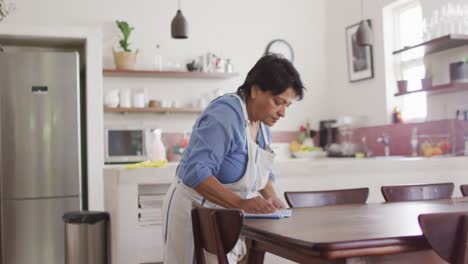 mujer biracial mayor con delantal y mesa de limpieza en la cocina sola
