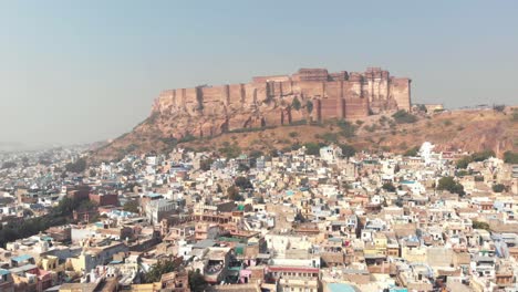 blue city landscape of jodhpur surrounding mehrangarh fort standing above cliff in rajasthan, india - aerial pan reveal shot