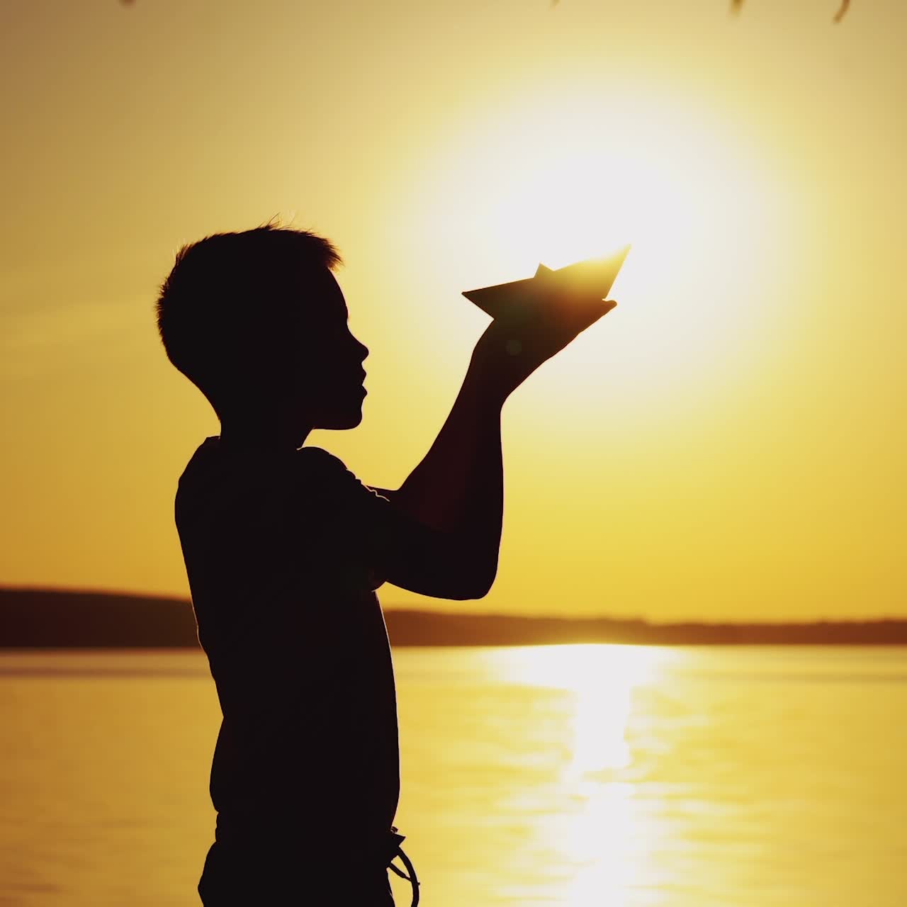 A little boy is holding an origami in the form of a paper boat in his hands and raising up it to the level of the sun on the background of the lake. Silhouette of a guy at sunset