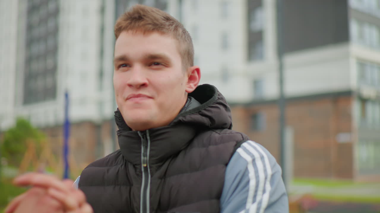 Ung, hvid mand jubler udendørs i boliggård med højhuslejligheder, iført puffer vest og hættetrøje, armene hævet og derefter foldet i et smil, nærbillede viser reflekterende, selvsikker