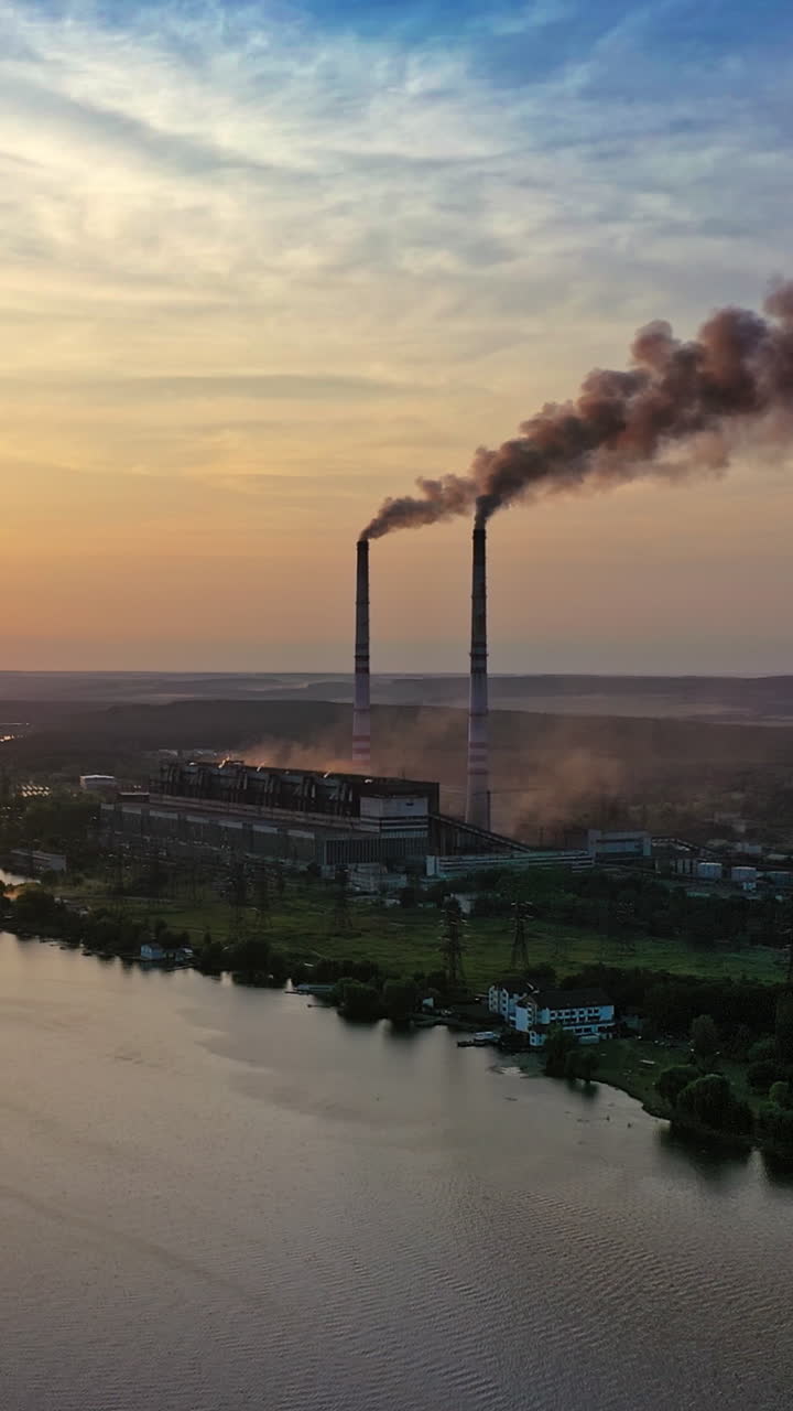 Dark smoke from industrial pipes near the water surface at sunset. Natural evening landscape of beautiful river and a factory. Pollution of the environment. Vertical video
