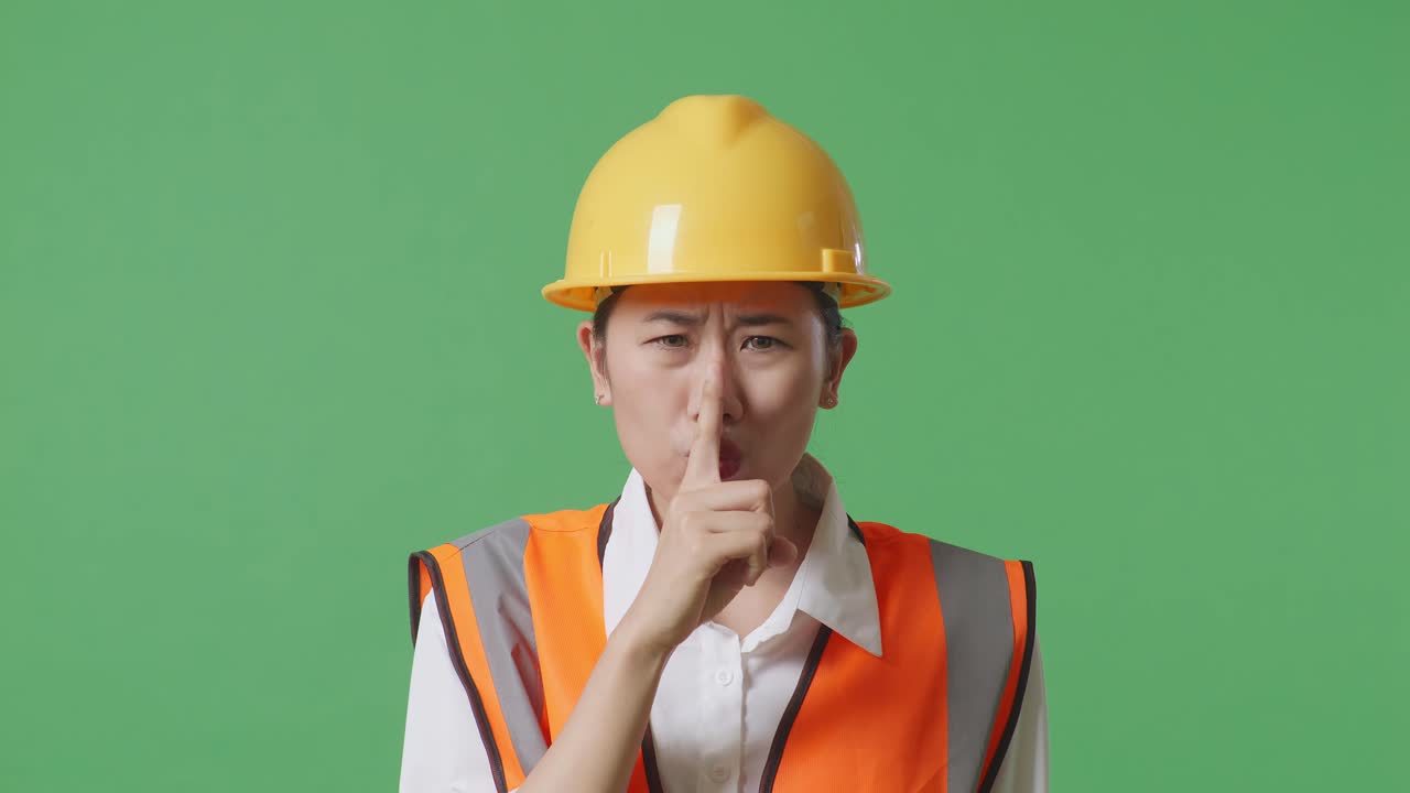 안전 헬을 입은 아시아 여성 엔지니어가 녹색 스크린 배경 스튜디오에 서서 shh 제스처를 하고 있습니다.