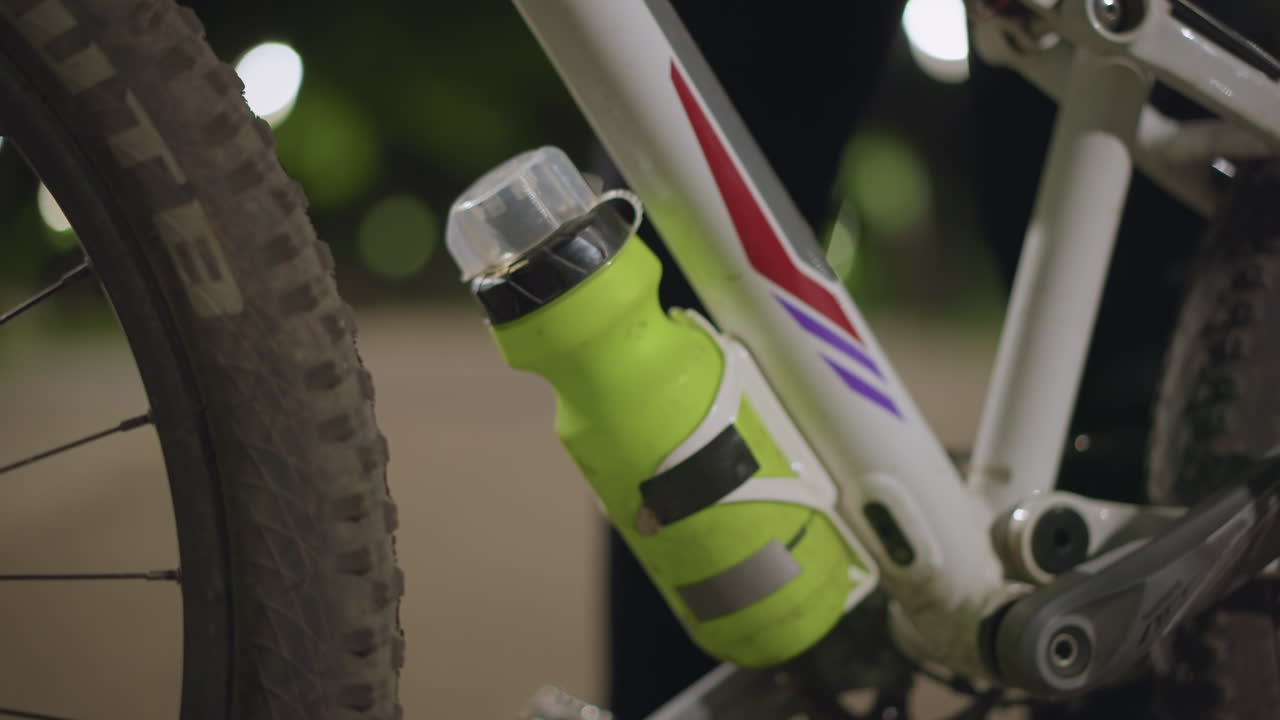 Green Bottle Clipped To Bicycle Frame Near Wheel, Close Detail Of Bottle Holder And Cap, Reflective Highlights From Park Lights, Hydration Accessory Ready For Night Ride Or Training