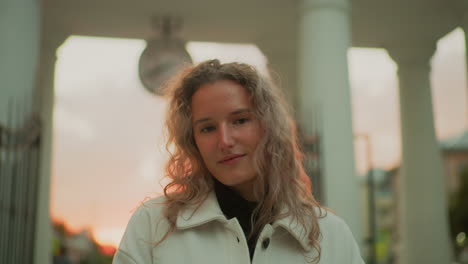 close up of woman standing with head slightly tilted down wearing white coat in soft sunset light, background showing classical pillars and city buildings