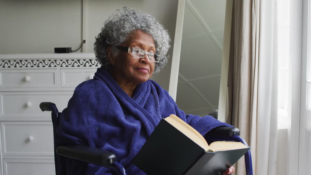 una mujer afroamericana mayor sentada en la silla de ruedas leyendo un libro en casa
