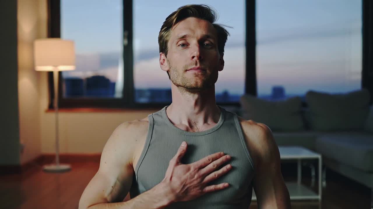 Uomo che fa stretching con canotta grigia, piegandosi, sollevandosi e centrando il respiro sul tappetino da yoga di casa, con il crepuscolo che avanza.