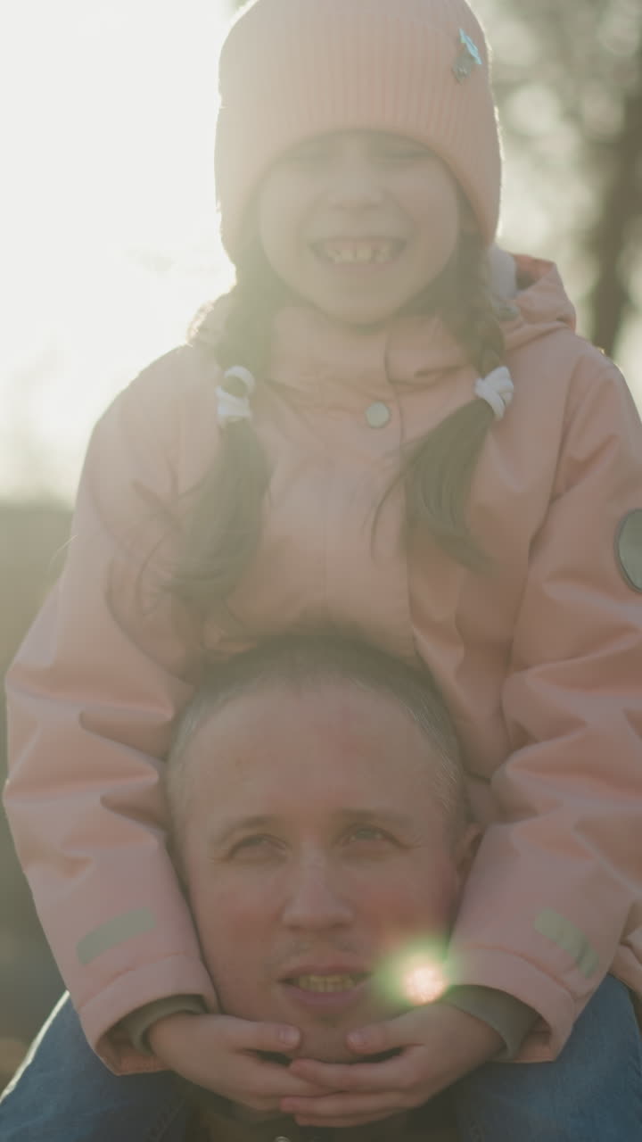 vista anteriore di una ragazzina con un berretto e una giacca rosa seduta gioiosamente sul collo di suo padre mentre camminano attraverso un parco illuminato dal sole. la ragazza sorride con gli occhi chiusi