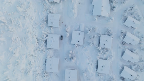 Toma-De-Drones-Con-Vista-Superior-Siguiendo-Un-Automóvil-Conduciendo-En-Medio-De-Cabañas-Nevadas-En-Laponia