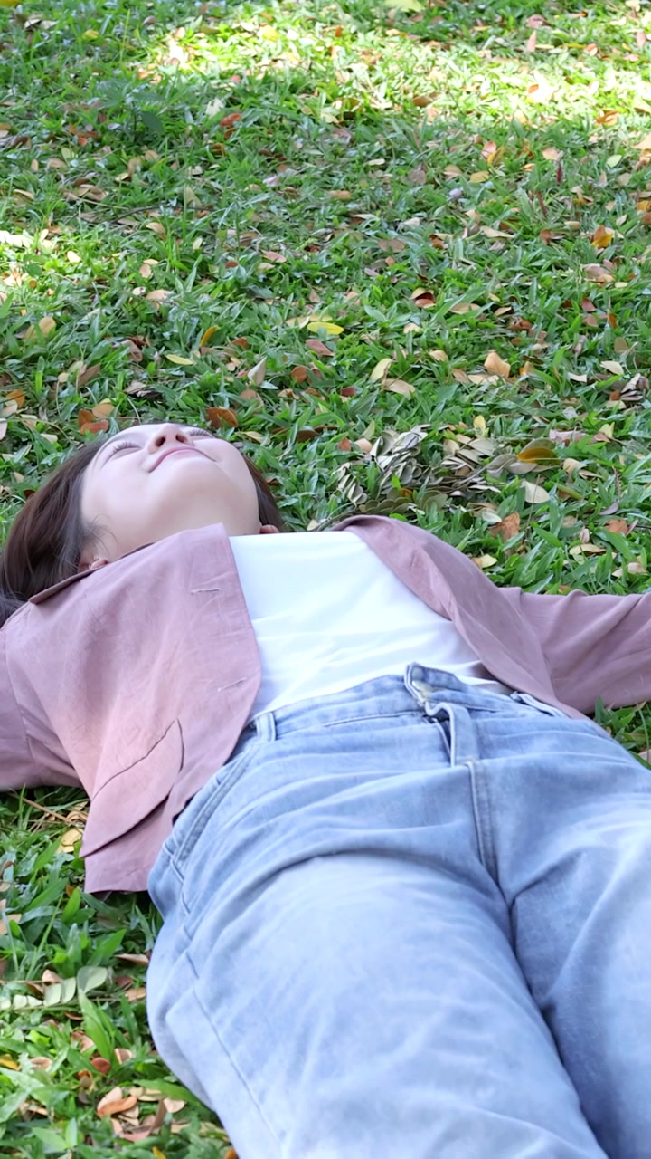 A smiling Thai university student lies on green grass in a sunny outdoor park