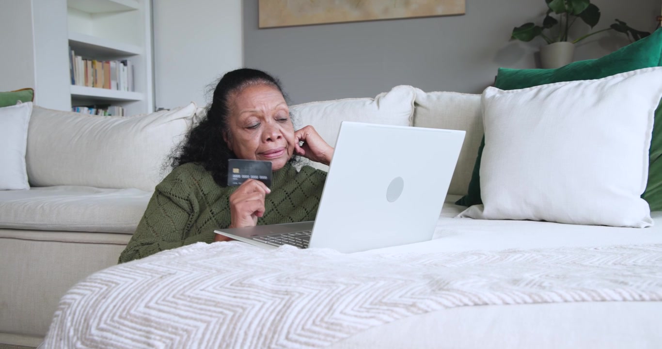 Senior African American woman checking bill on laptop at home holding card, calling, closing laptop