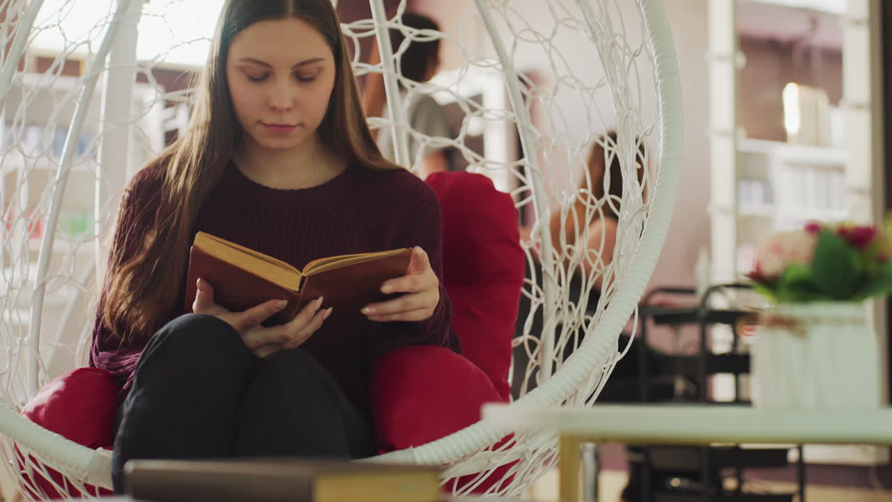 Mujer caucásica disfrutando de un momento de lectura tranquilo, joven sentada cómodamente en un ambiente de lectura acogedor, estudiante sumergida en un libro en un salón tranquilo con luz cálida del sol