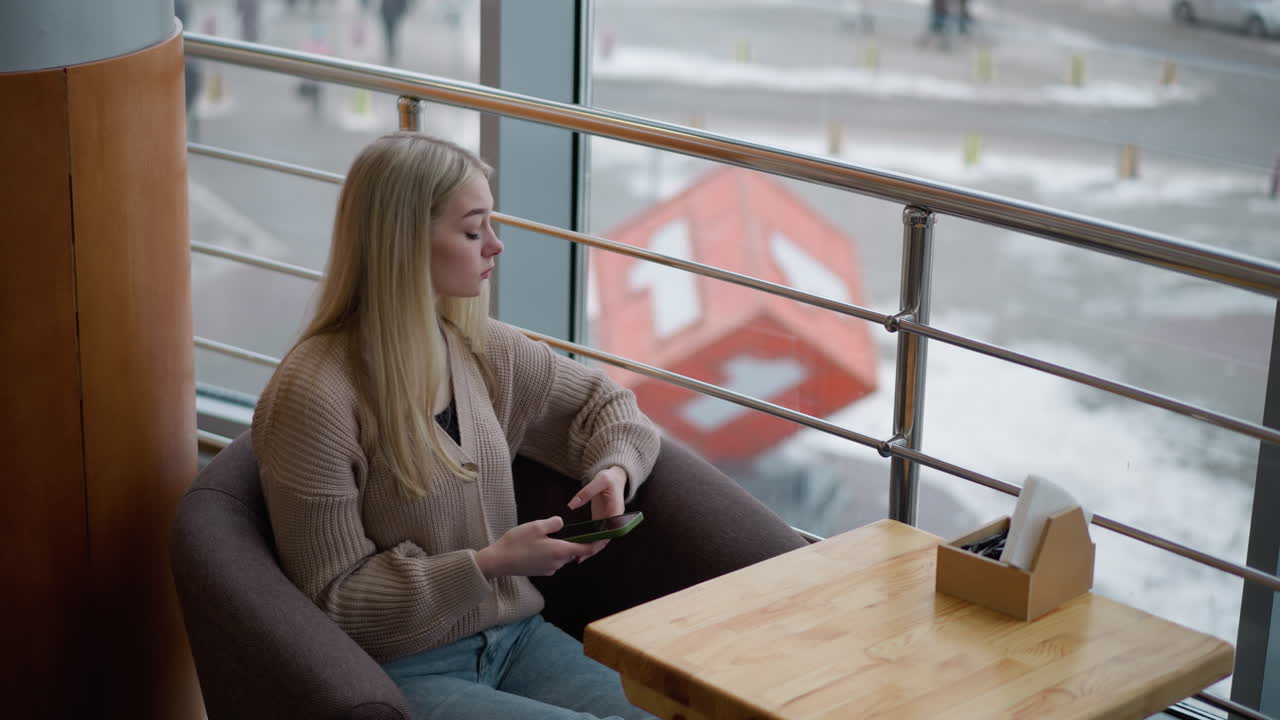 mujer elegante sentada en un restaurante, sosteniendo el teléfono y mirando pensativo, esperando a alguien, vista de la ciudad a través de una gran ventana de vidrio con nieve y escena urbana en el fondo