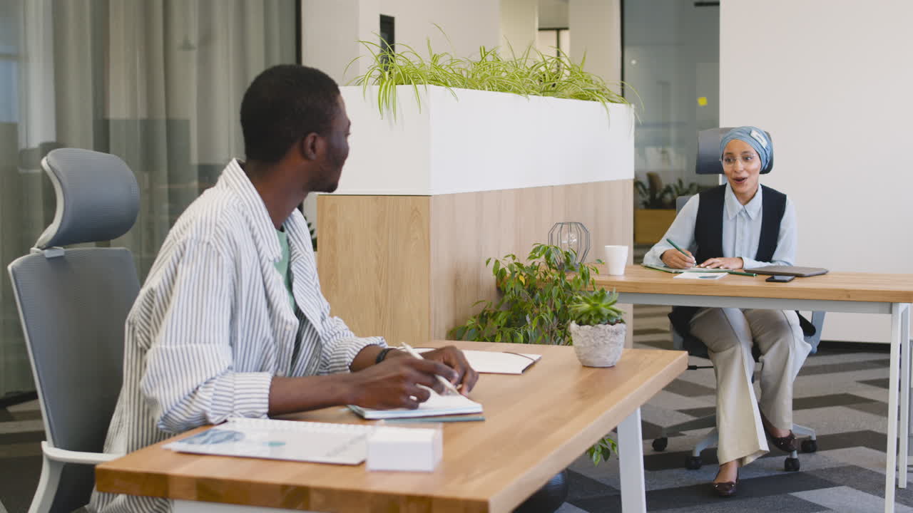 Young Smiling Worker Working Taking Notes Sitting At His Desk And Talking With Muslim Businesswoman Who Works Sitting At Their Desk