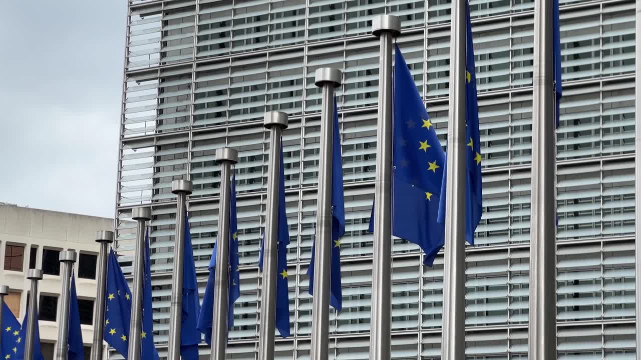 Molteplici bandiere dell'Unione Europea che sventolano al vento di fronte a un moderno edificio vetrato adibito a uffici a Bruxelles