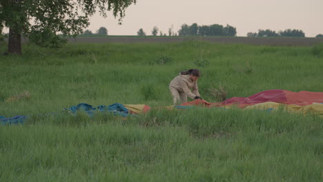 woman pulling red hot air balloon fabric through tall grass at sunset with gloves while pilot assists from wicker basket next to trailer during preflight pack up in rural open landscape scene