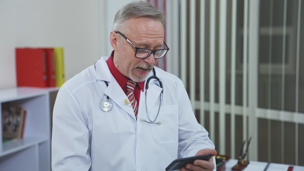 Medico anziano seduto alla scrivania del medico e che guarda il telefono. Medico esperto e pensieroso consulta il paziente tramite videoconsulto.