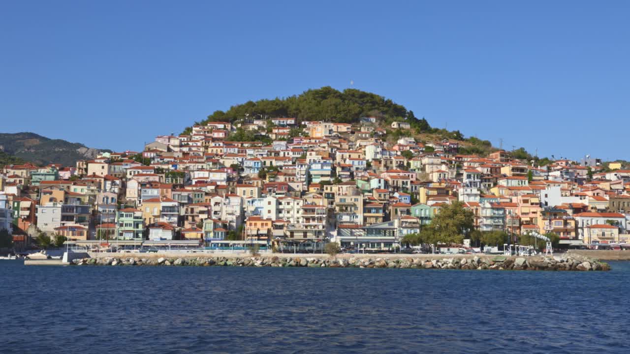 A panoramic view of a Greek town with terracotta rooftops and hills in the background.