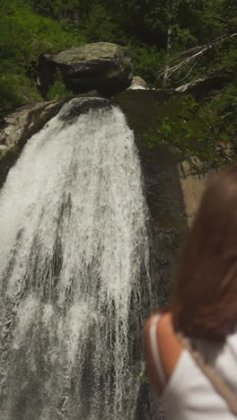 foamy waterfall sprays on steep cliff and mother with little son rests in wild park. woman shows whitewater cascade to toddler boy on weekend hike