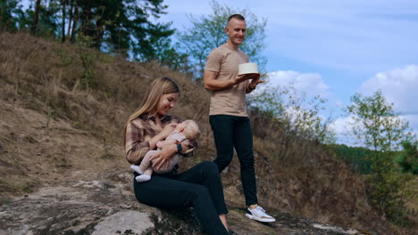 long-haired caucasian woman sits on the rock with her baby son in hands. man with a cake goes down the hill to join them.