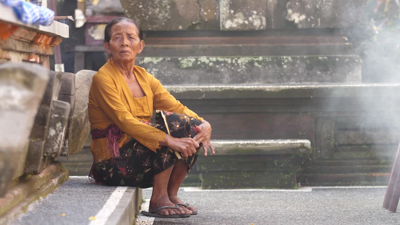 Elderly woman sits on stone step in Hindu temple on island of Bali, Indonesia with smoke from fire and incense fills scene