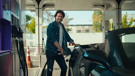 A-happy-brunette-guy-in-a-denim-jacket-refuels-a-gray-convertible-and-looks-at-the-camera.-Gray-cabriolet-owner-posing-and