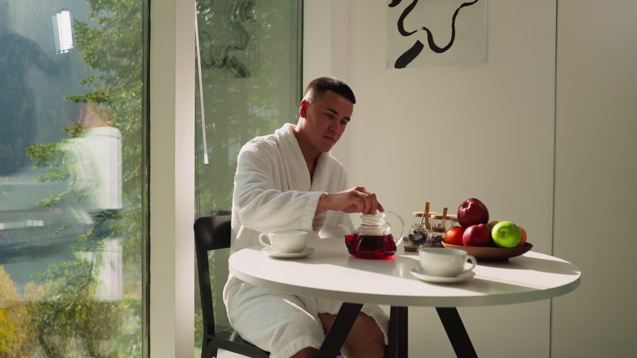 주방에 앉아있는 차병에 을 는 남자: 색 욕조를 입은 예쁜 젊은 신사는 파노라마 창문 근처의 테이블에서 차 의식을 즐긴다.