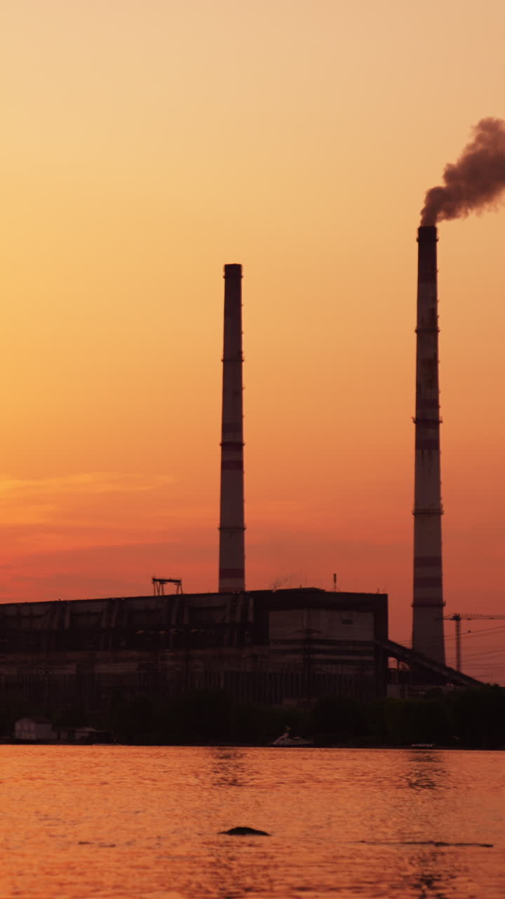 Bright setting sun hiding in the clouds at sunset. Industrial plant working near the river producing thick smoke from high pipe. Vertical video