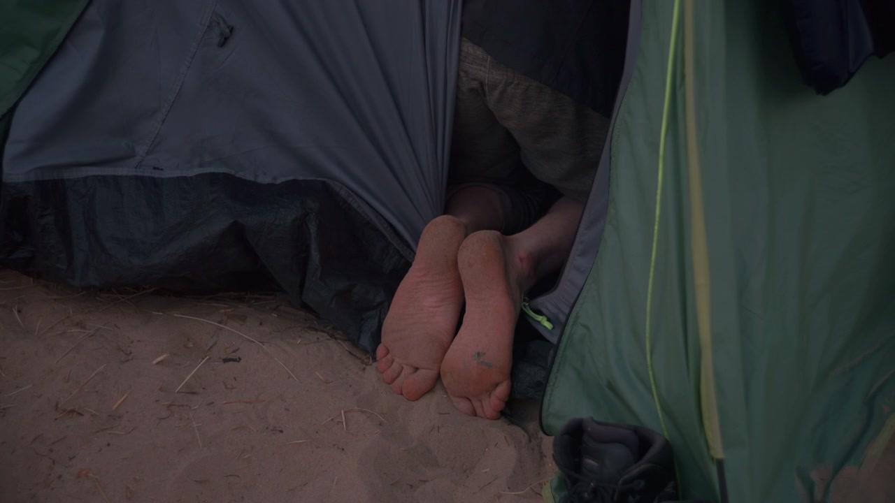Pies que sobresalen de debajo de la solapa de una tienda de campaña sobre la arena, capturando un momento relajado de descanso durante un viaje de acampada en la playa