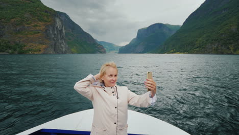 a happy woman takes pictures of herself on the nose of a cruise ship sails through a picturesque fjo