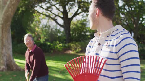 Video-portrait-of-happy-caucasian-adult-son-raking-up-autumn-leaves-with-senior-father-in-garden