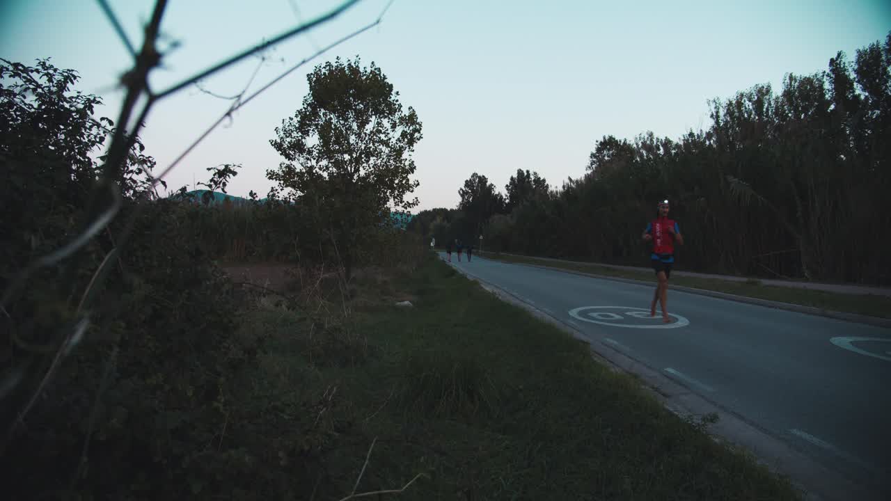A runner runs barefoot on the road, early in the morning as the sun rises, with a light on the head due to the low light. View from behind a plant