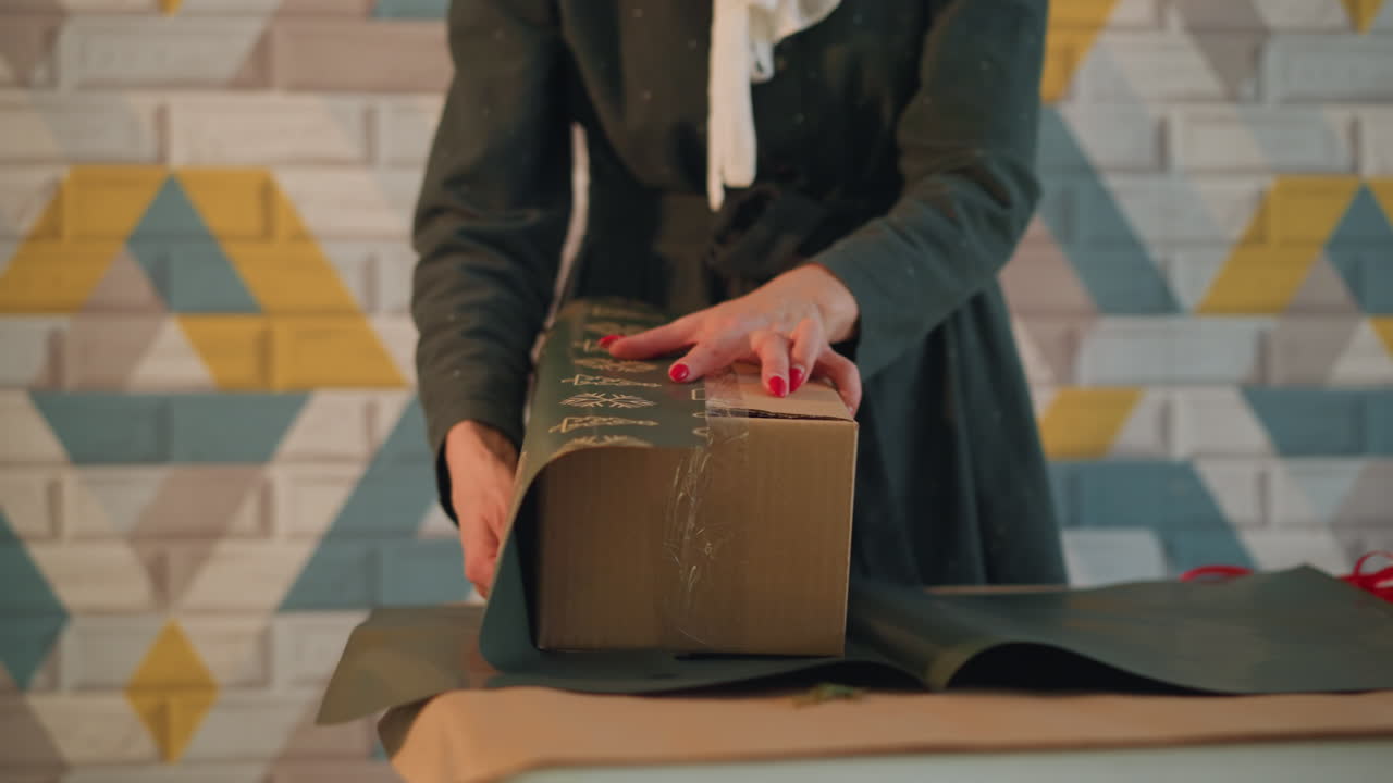 Mani che impacchettano un regalo di Natale su un tavolo con scatola marrone, nastro rosso e ornamenti dorati; la donna piega con cura la carta, applica il nastro adesivo e leviga i bordi in uno studio caldo con sfondo a parete geometrica e luce soffusa.