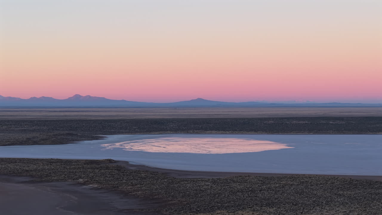 Drone ascends over the vast salt flats of Salinas del Diamante in Mendoza, Argentina, revealing reflective surfaces, arid terrain, and the distant snow-capped Andes under a pale blue sky