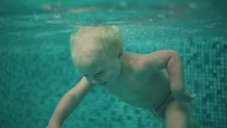 cute blonde toddler is diving under the water in the swimming pool and swimming there until his mother is lifting him from the water. an underwater shot