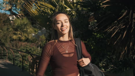 Medium-shot-of-charming-woman-with-football-and-backpack-walking-along-city-park-after-training.