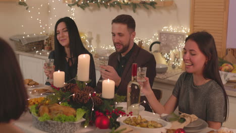 group of happy friends sitting at table, holding champagne glasses and talking to each other at home on christmas 1