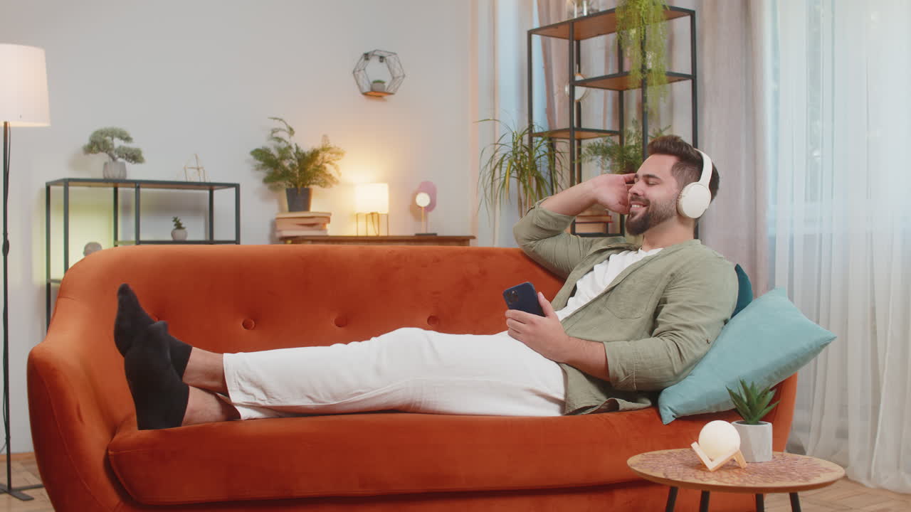joven feliz y encantado con auriculares inalámbricos usando un teléfono inteligente relajándose sentado en el sofá en casa