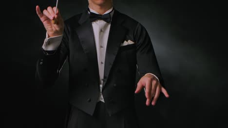 concert of classical music under direction of an orchestra conductor. man waves his baton to synchronize and conducting musicians in theater. black background with smoke and backlight. close up