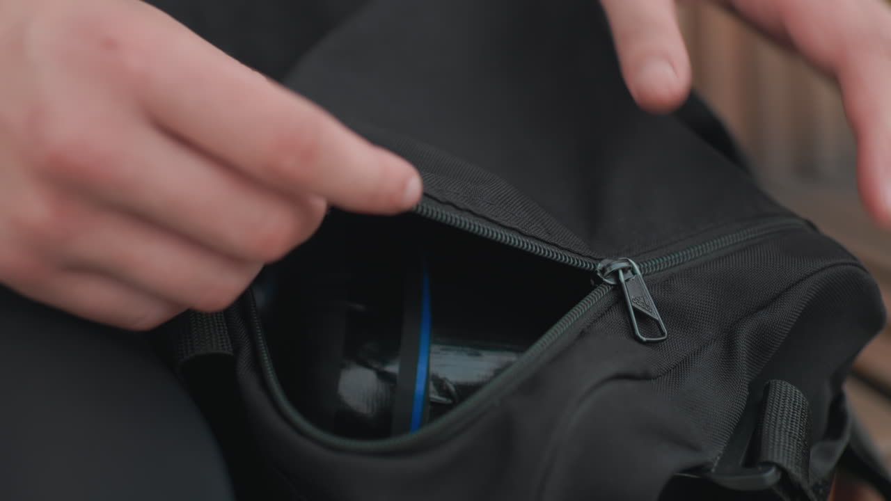 Manos abriendo una mochila negra para revelar un pequeño objeto azul metido en el bolsillo delantero; los primeros planos muestran una búsqueda cuidadosa, detalles de la cremallera y ajuste de la correa; banco urbano de fondo, ambiente de camino al trabajo por la mañana.
