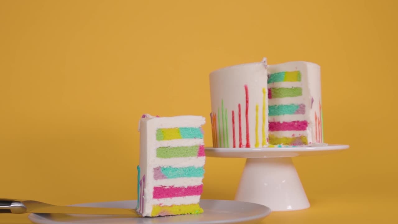 A vibrant slice of rainbow cake sits on a plate against a solid yellow backdrop