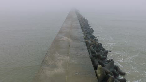 aerial establishing view of port of liepaja concrete pier, baltic sea coastline , foggy day with dense mist, moody feeling, big storm waves splashing, birdseye drone shot moving forward low