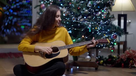 wide shot of confident cheerful middle eastern young woman playing guitar singing sitting at decorated christmas tree on new year's eve. portrait of talented female musician celebrating holiday at home.