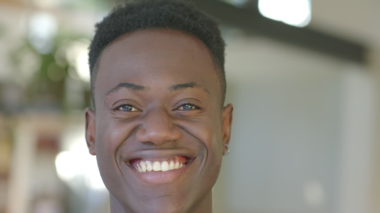 Un homme au visage neutre, souriant et riant sur le plateau, réagissant à une blague hors champ, portant une boucle d'oreille créole.